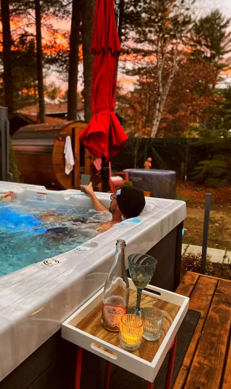 Personne dans le spa au coucher du soleil avec un plateau de vin et le sauna derrière